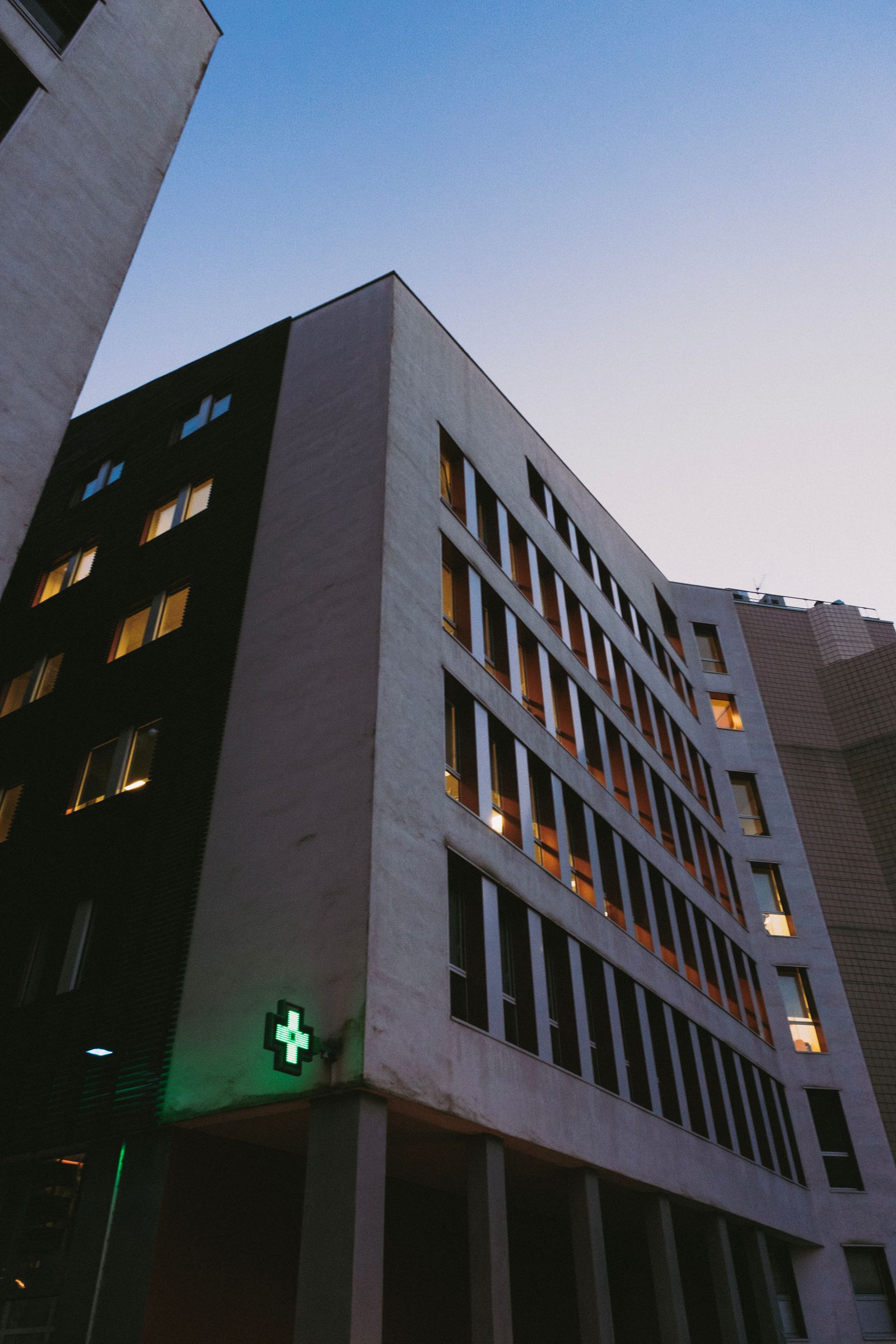 Façade d'un bâtiment hospitalier au crépuscule, avec une croix verte lumineuse.